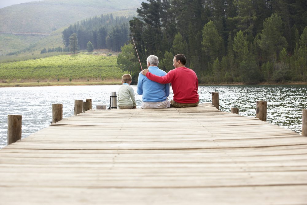 A Father, Son and Grandfather sitting on the edge of a pier fishing on a lake.